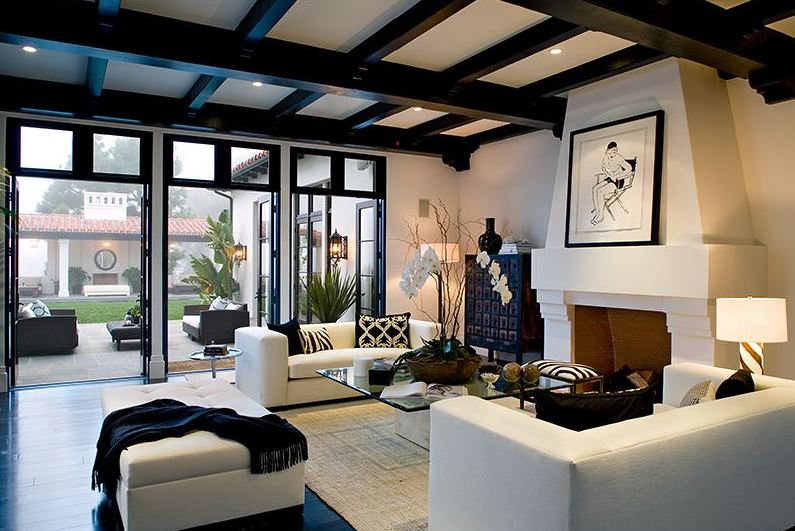 Living room in a Spanish revival home with exposed painted black beams, a grand fireplace, a jute rug, two white sofas and a large tufted ottoman 