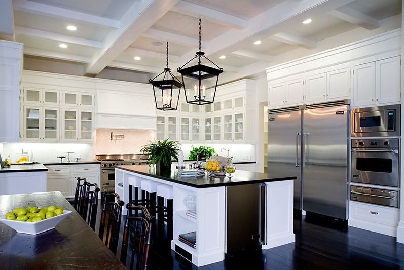 Eat in kitchen in a Spanish revival home with two lantern lights, white cabinets with glass fronts, coffered ceiling, stainless appliances and a white island with black countertops