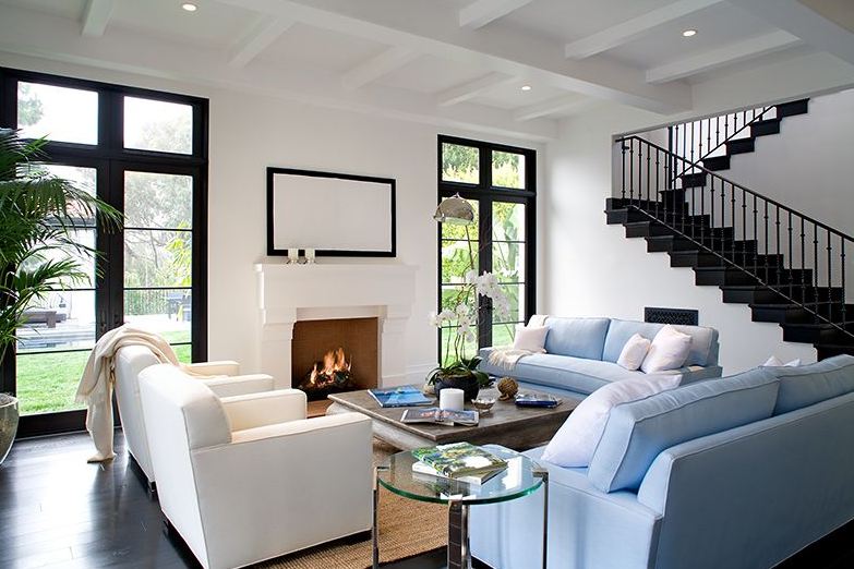 Living room in a Spanish revival home with light blue sofas, white armchairs, dark wood floor, black glass doors and a white fireplace