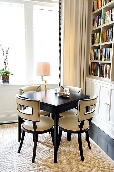 Side table in a living room surrounded by dark wood chairs with white leather seats and back with nail head trim, floor length curtains and a white built in bookshelf