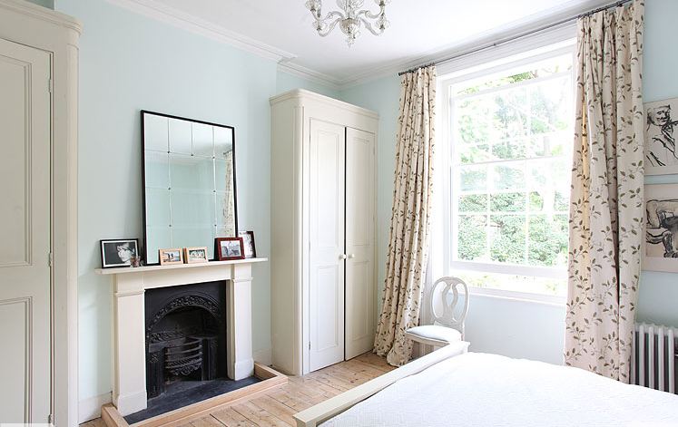 White bedroom in a London home with tall cabinets, fireplace, wide wood plank floor and leaf print curtains 