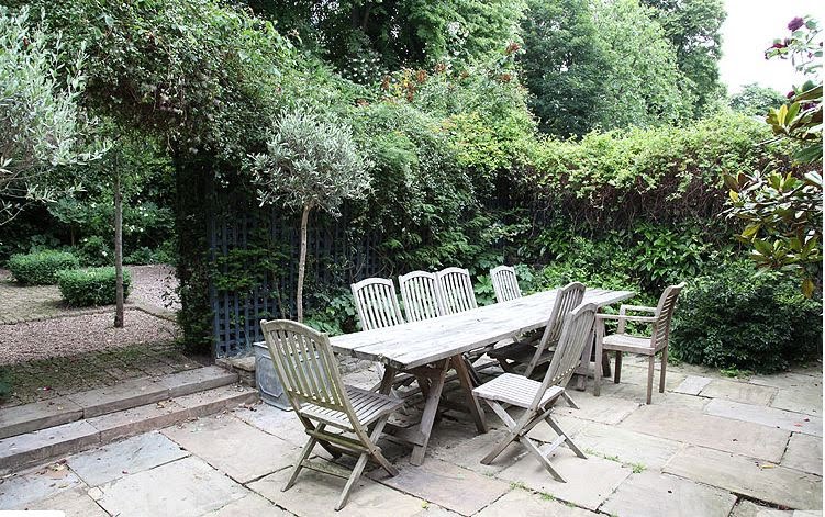 Long teak outdoor dining table sits in the paved part of the garden near the kitchen