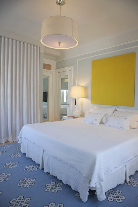 White bedroom with a blue rug and large yellow square above the bed instead of a headboard