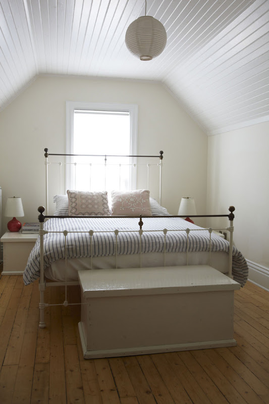White bedroom with white beadboard ceiling, wide plank wood floor, white iron bed frame and a white lantern
