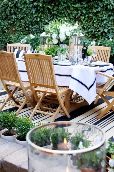 Outdoor dining party with blue and white striped rug, wood patio chairs and a blue and white table setting surrounded by a row of tiny boxwood trees and a wall of ivy