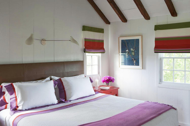 Bedroom in the Hamptons with leather headboard and white bedding with purple and red stripes and Roman shades