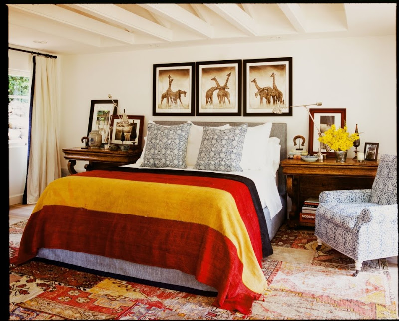 Bedroom by Martyn Lawrence Bullard with patterned area rug, striped throw, blue and white patterned armchair, and three framed pictures of giraffes over the headboard