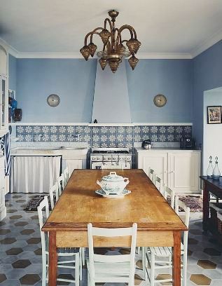 Blue kitchen in an Italian villa with hexagon tile floor, long wood table surrounded by white chairs, blue and white tile backsplash and a bronze chanedlier