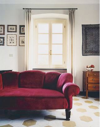 Living room in an Italian villa with a red velvet sofa, gold and white tile floor, framed prints, a wood chest of drawers and a window