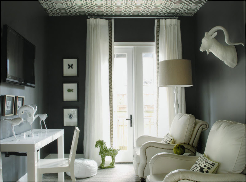 Dark grey TV room with large glass door with long sheer curtains, wallpapered ceiling, white leather armchairs with nail head trim, a white desk, wall mounted TV and a white ram head