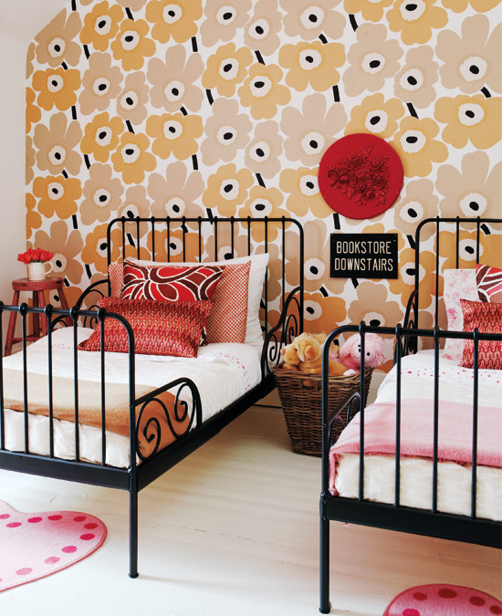 Bedroom with Marimekko wallpaper, two twin beds with black iron frames and white bedding with brightly colored pillows, and a light wood flor