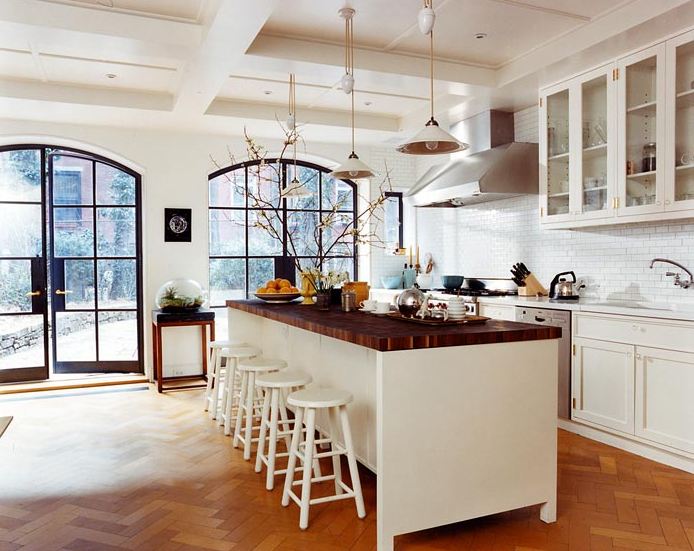 White open kitchen with butcher block countertop, herringbone wood floor, huge arched multi-paned glass doors and window, two white pendant lights, white subway tile backplash, white drawers and cabinets and a white island surrounded by white stools