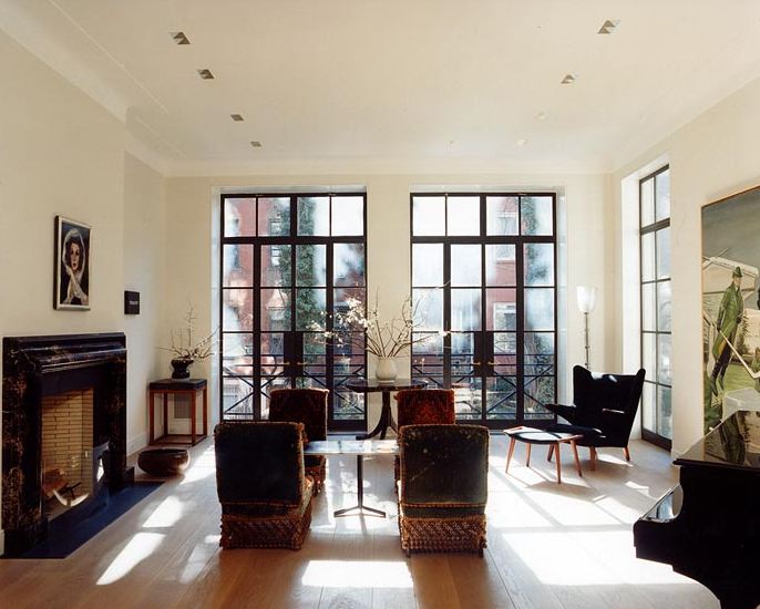 Living room in a New York townhouse with three floor to ceiling paned windows and doors, wood floor, marble fireplace, dueling armchairs, and a grand piano