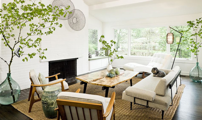Living room with white painted brick fireplace, reclaimed wood coffee table, sisal rug,wood armchairs, white chaise lounge, taupe sofa. and large glass jars holding tree branches
