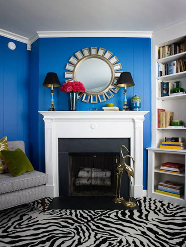 Living room with royal blue walls, grey sofa, white fireplace with a sunburst mirror on the mantel, built in bookshelves and zebra print carpeting