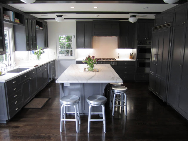 Kitchen with dark grey cabinets and drawers, marble counter tops, wood floor, and metal barstools around a large island with a marble top