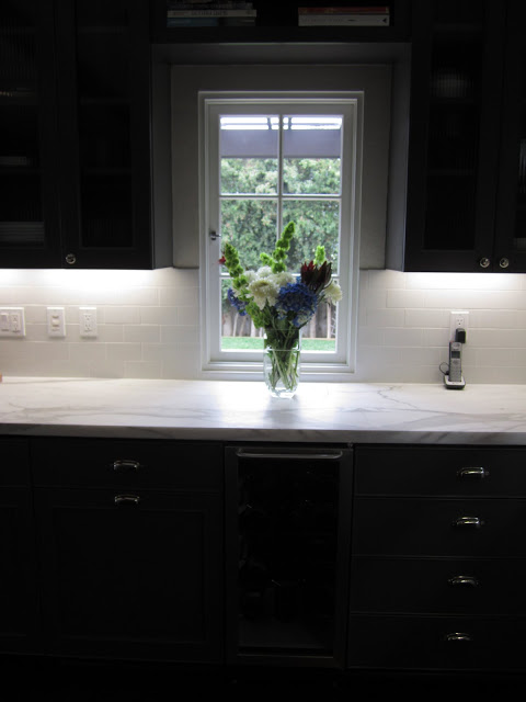 Kitchen with grey cabinets and drawers, marble counter top, casement window and a flower arragement