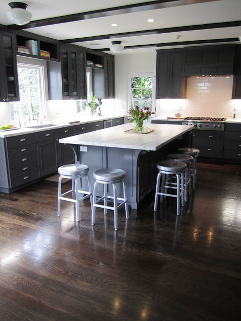 Kitchen with dark grey cabinets and drawers, school house lighting and dark stained wood beams, marble counter tops, wood floor, and metal barstools around a large island with a marble top