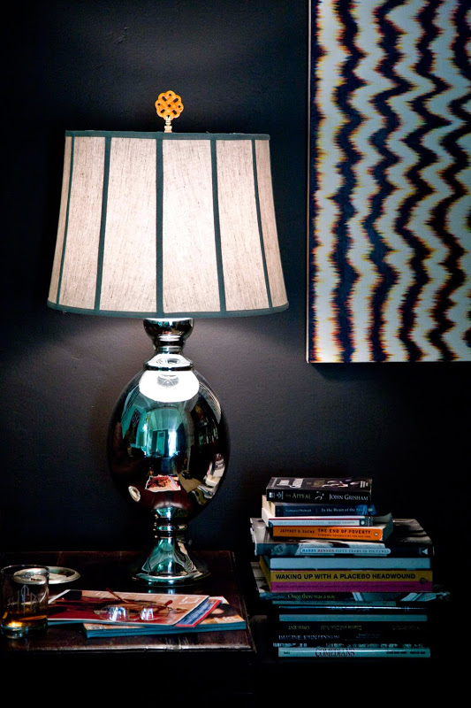 Side table with a polished metal lamp with a gold finial from Hillary Thomas Designs, a stack of books in a room with dark walls and a framed ikat print on the wall