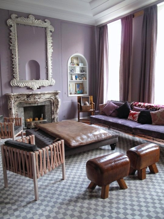 Purple living room in a Brussels apartment with lavender walls, grey and white checkerboard floor, antique fireplace with a large white mirror on the mantel, tufted black ottoman, to leather stools, armchairs with leather straps and a purple sofa with exposed metal legs