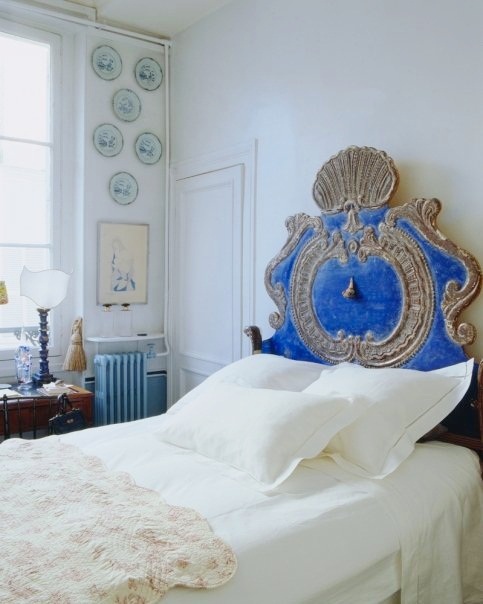 White bedroom in a Paris Apartment with decorative plates on the wall and an 18 century metal and blue Venetian headboard