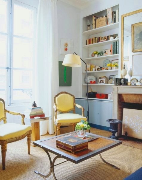 Living room in a Paris apartment with yellow Louis XIV chairs, a sisal rug on a wood floor, a coffee table with metal legs and a wood top, fireplace and built in bookshelves