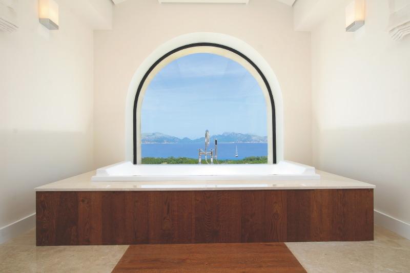 Bathroom with arched picture window with a view of the water, massive stand alone tub, a wood walkway to the tub and marble floor