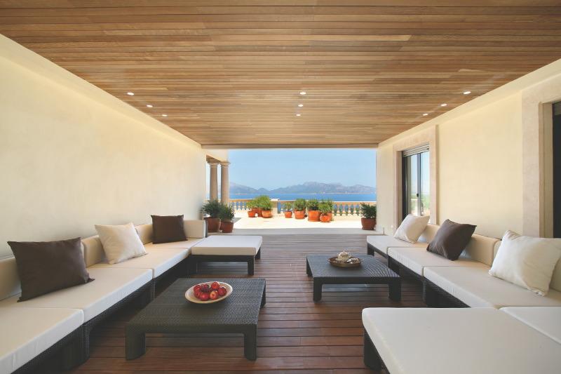 Covered backyard patio with sectional sofa style lounge chairs, wood floor, woven ottomans, and a view of the water
