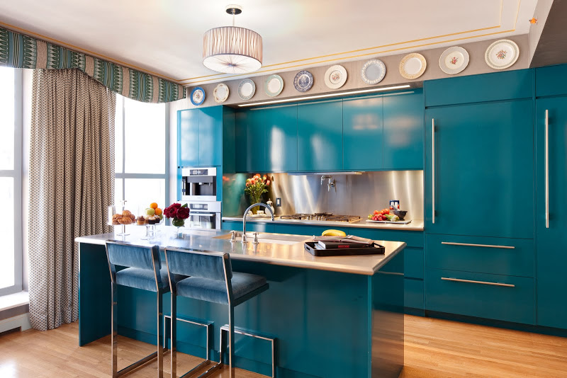 Blue kitchen that Zac Posen collaborated on with a wood floor, cabinets and drawers with long silver pulls, decorative plates, a drum pendant light, large windows with grey floor length curtains, at the island there are two bar chairs with blue velvet seats and backs