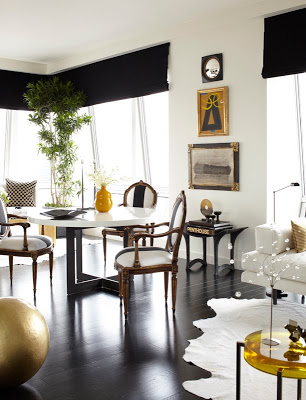Dining room in an NYC home with dark wood floor, white table with dark legs, upholstered Louis XIV chairs, black roll up curtains, a white rug with a white sofa peaking in