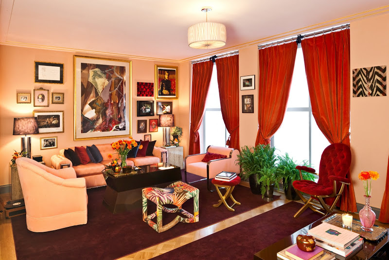 Living room that Zac Posen and Morris Adjmi collaborated on with Parquet wood floor, purple area rugs, large windows with orange curtains, peach walls, armchairs and sofa, a dark coffee table, a tufted red velvet seat with matching ottoman