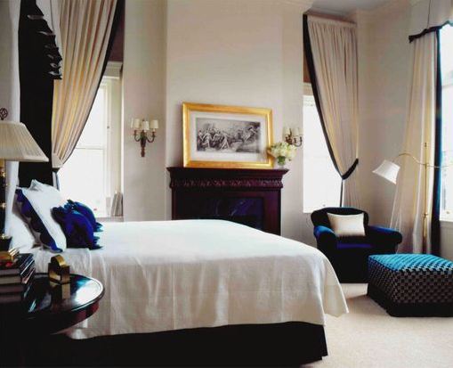 Bedroom with a fireplace, three windows with long white curtains with blue ties, a blue velvet armchair and woven ottoman, the bed has a large black headboard and the bedding has blue trim 
