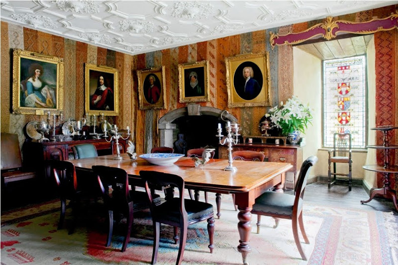 Dining room in Glin Castle with carved ceiling, brightly colored wallpaper, a fireplace, portraits in gold grams, a wood table and chairs with blue-grey seats and back, a stained glass window with family crests and a large rug