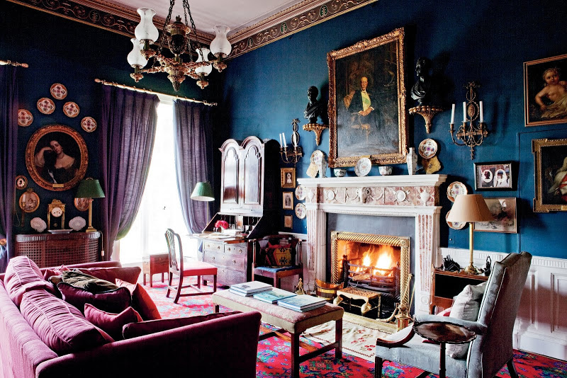 Dark blue library with a fireplace, decorative ceiling molding, burgandy soffa, writing desk, portraits in traditional frames, purple curtains, a gold chandelier and red patterend carpet in Glin Castle in Ireland