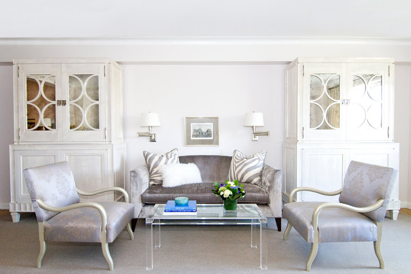 Elegant, kid friendly living room in NYC with lavender armchair and sofa, a lucite coffee table, light grey carpeting, two wall mounted lights and two large white cabinets