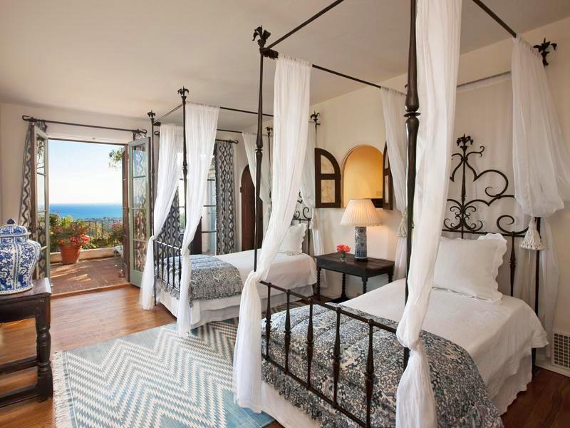 Bedroom in a Montecito mansion with wood floor, blue and white chevron printed rug, two iron canopy beds with white bedding and a brocade throw at the foot of the bed, and double doors with blue and white curtains lead to an outdoor patio