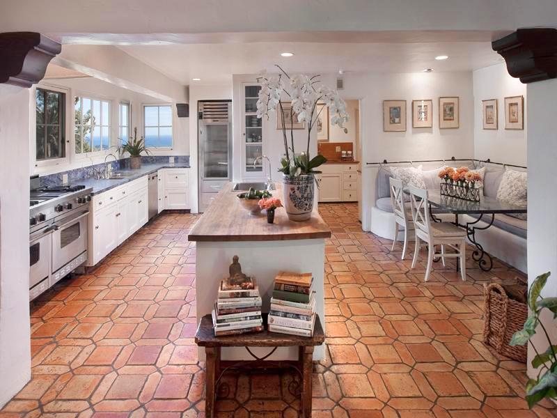 Kitchen in a Montecito mansion with stainless appliances, tile floor, columns, white drawers and cabinets, dark counter tops, an island with a wood counter top and sink and a breakfast nook with banquette seating and a glass table with metal legs 