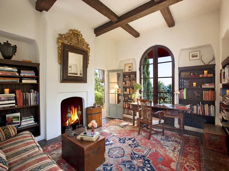 Library in a Montecito mansion with a fireplace and a mirror in a traditional frame on the mantel, built in bookshelves, arched windows, a wood desk and chair, a striped sofa and a large Turkish rug