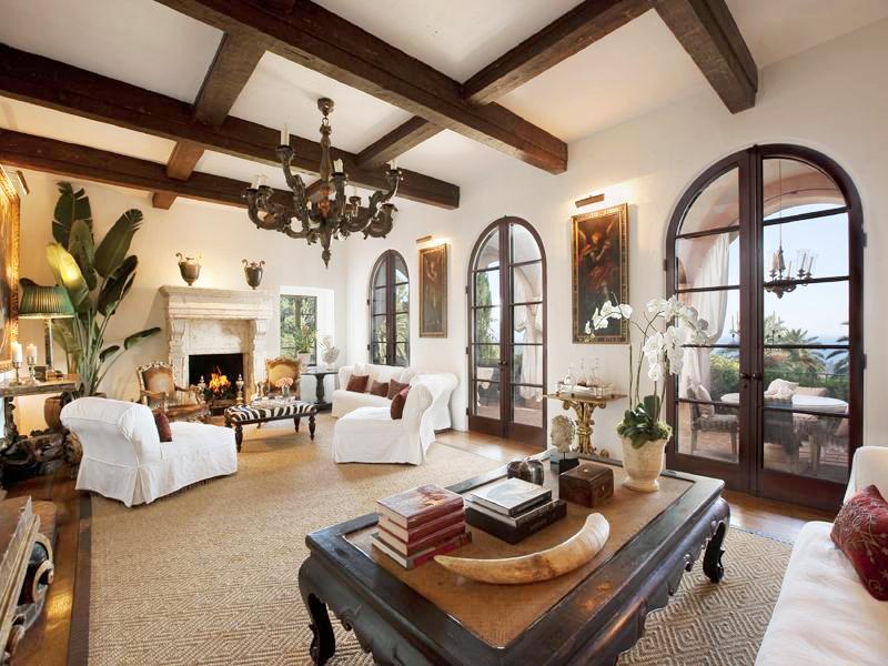 Living room in a Montecito mansion with exposed beams, wood floor, a tan rug, chandelier, white armchairs and sofa and high arched doors leading to a balcony