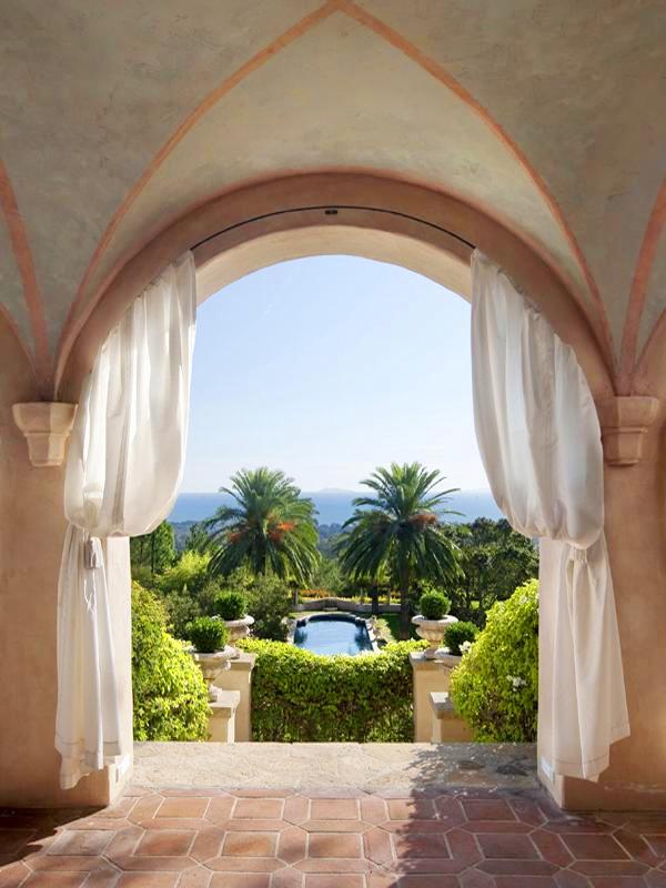 Arched entryway to the backyard of a Montecito mansion with white curtains tied back and saltillo tile floor