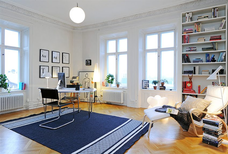 Alternative view of a home office in a Swedish apartment with  herringbone wood floor, carved crown molding, the decorative ceiling medallions, tall paneled doors, metal desk, and chair, a blue striped rug, built in bookcase and a white lounge chair with a stack of books, and a globe light