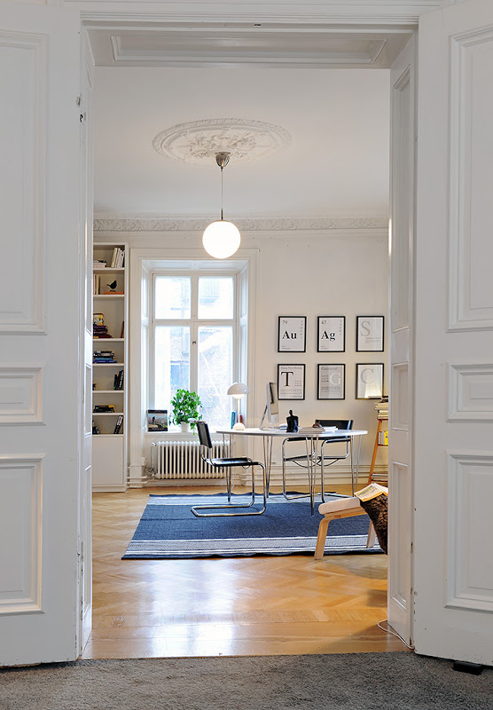 Home office in a Swedish apartment with  herringbone wood floor, carved crown molding, the decorative ceiling medallions, tall paneled doors, metal desk, and chair, a blue striped rug and a globe light