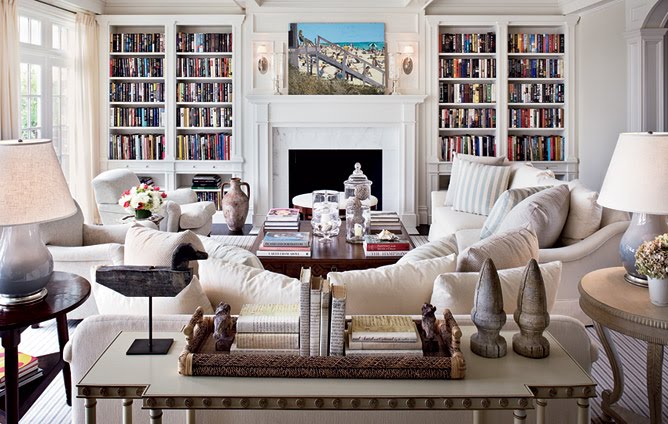White living room with two sofas, arms chairs, wood side tables, a striped rug, built in bookcases and a fireplace