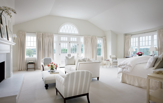White bedroom with french doors, large windows, a fireplace, sofa, grey and white striped dueling armchairs and an upholstered ottoman