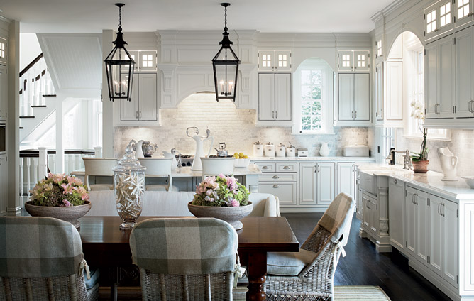 White kitchen with black lantern lights, wood floor, and a wood table surrounded by white wicker chairs with grey gingham covers