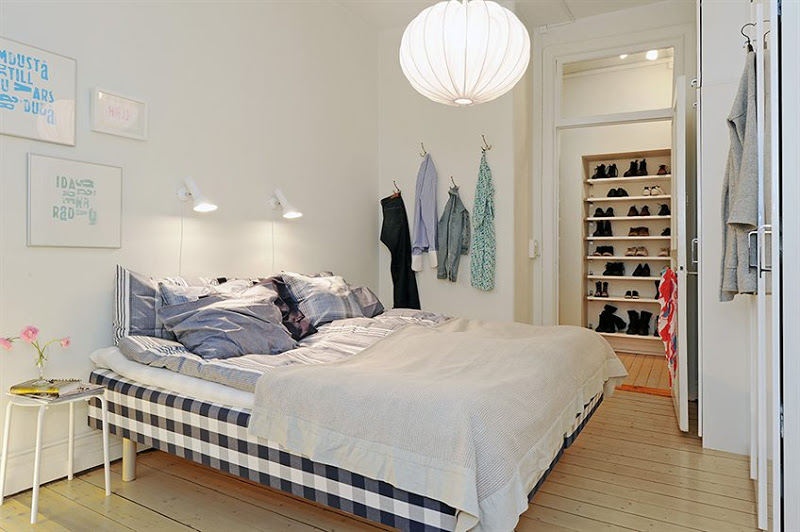 Bedroom with light wood floor, a gingham bed, wall mounted reading light, a bright white lantern and a walk in closet