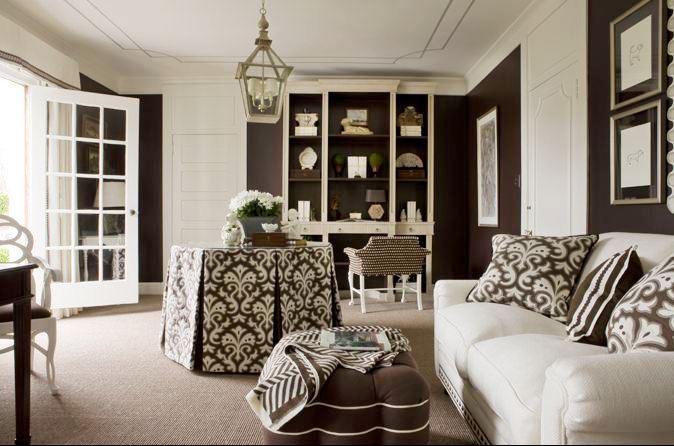 Sitting room with brown walls, a white sofa, tufted brown ottoman with white piping, a lantern, built in bookshelf and a round table with a brown and white table cloth