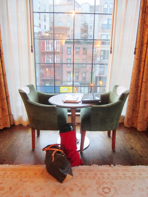 Sitting area in a suite at The Bowery Hotel with two green armchairs around a round table, a floor to ceiling window with a view, wood floor and patterned curtains