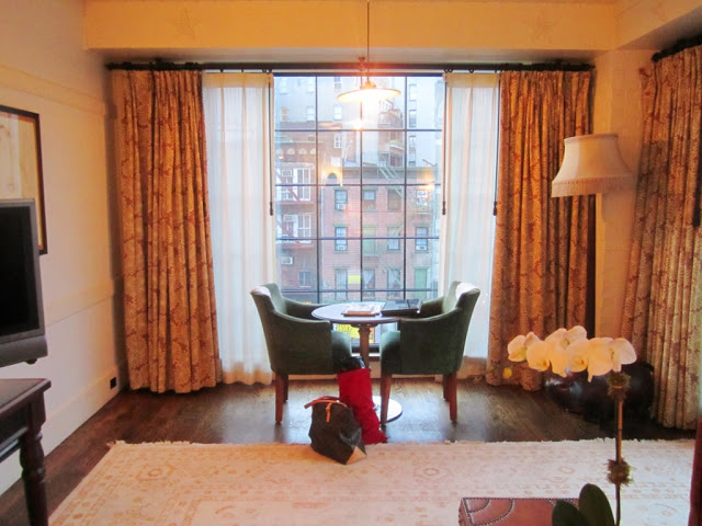 Sitting area in a suite at The Bowery Hotel with two green armchairs around a round table, a floor to ceiling window with a view, wood floor and patterned curtains