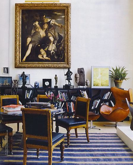 Living room with a round dark wood table, wood chairs with blue leather seats and backs and nail head trim, a blue and cream striped rug, black bookshelf, a tan Egg Chair with matching ottoman and a large classical painting in a gold frame